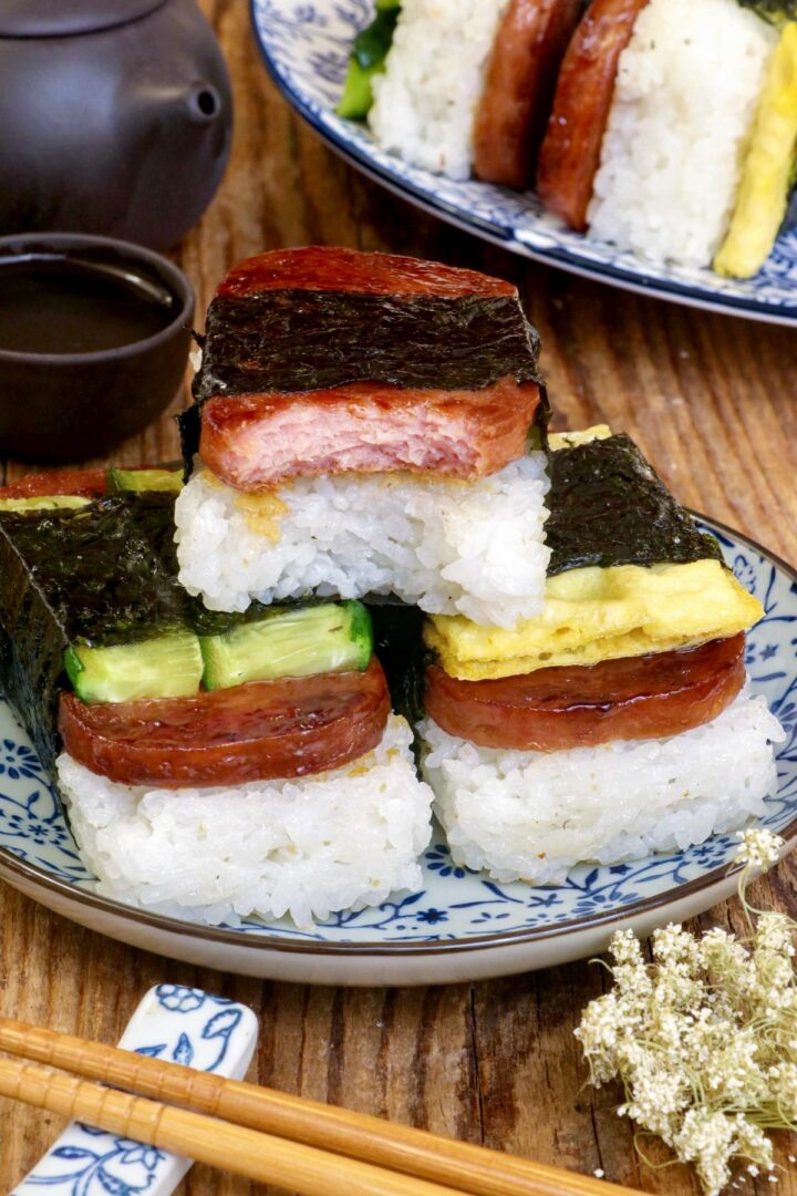 Variety of Spam Musubi with luncheon meat, cucumber, an egg roll on a serving plate.