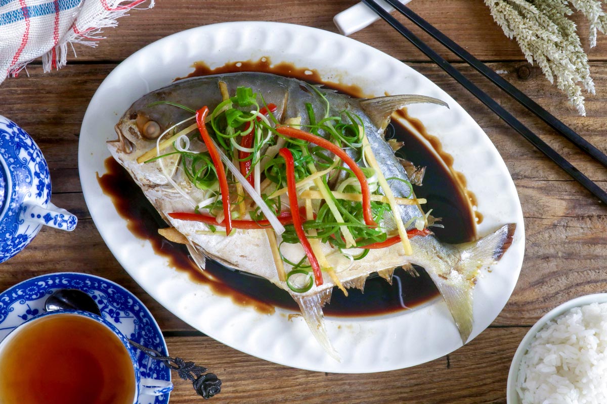 Steamed pompano fish Chinese- style in a serving platter with sliced ginger, green onions, and cilantro on top.